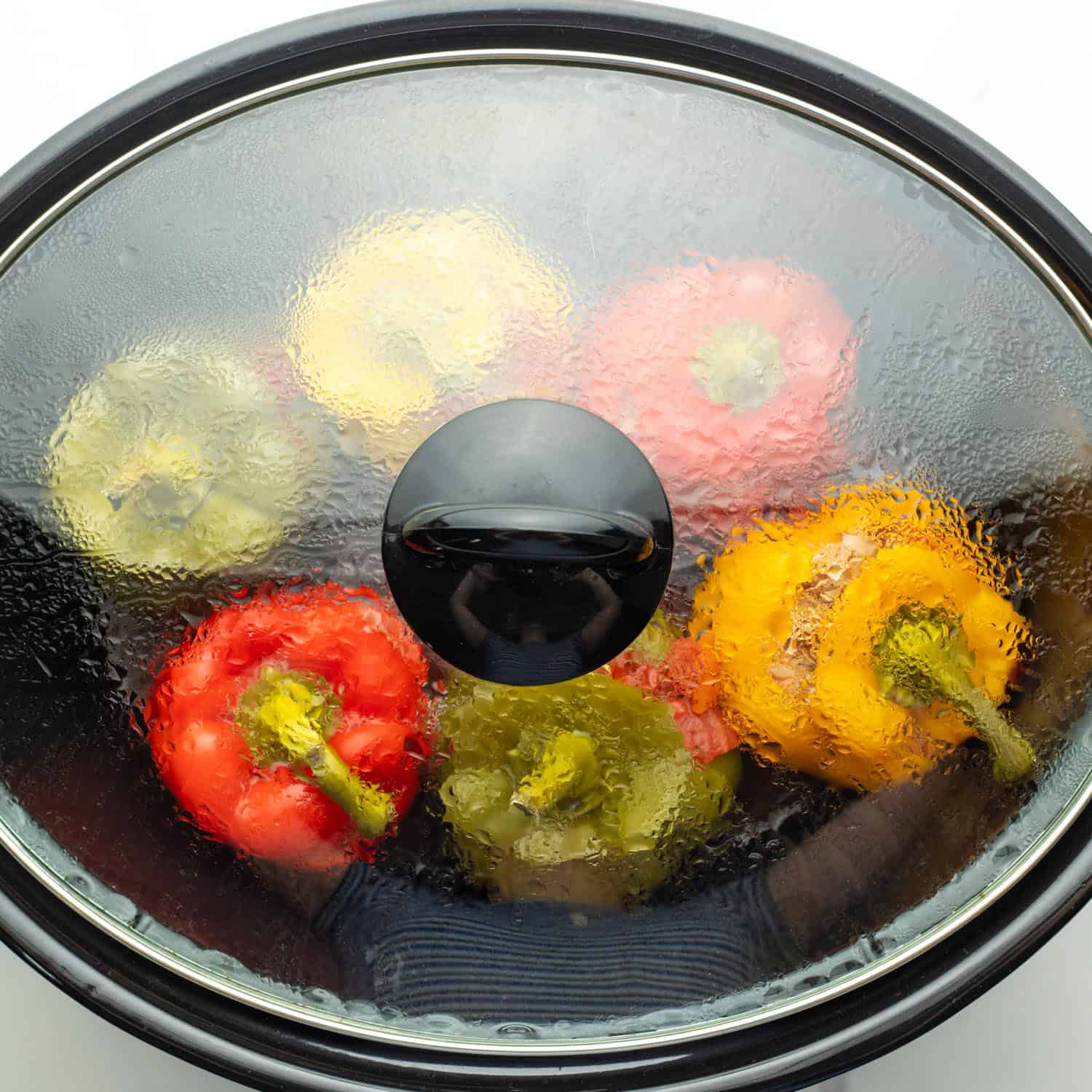 A covered slow cooker holding 6 large stuffed bell peppers in various colors.