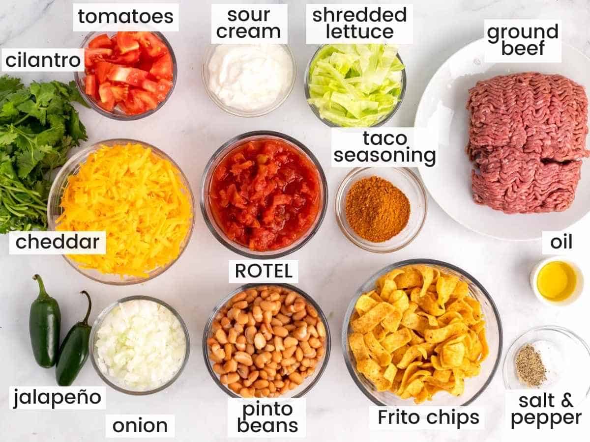 Ingredients needed to make a walking taco casserole including ground beef, cheese, frito chips, pinto beans, lettuce, seasonings and aromatics.