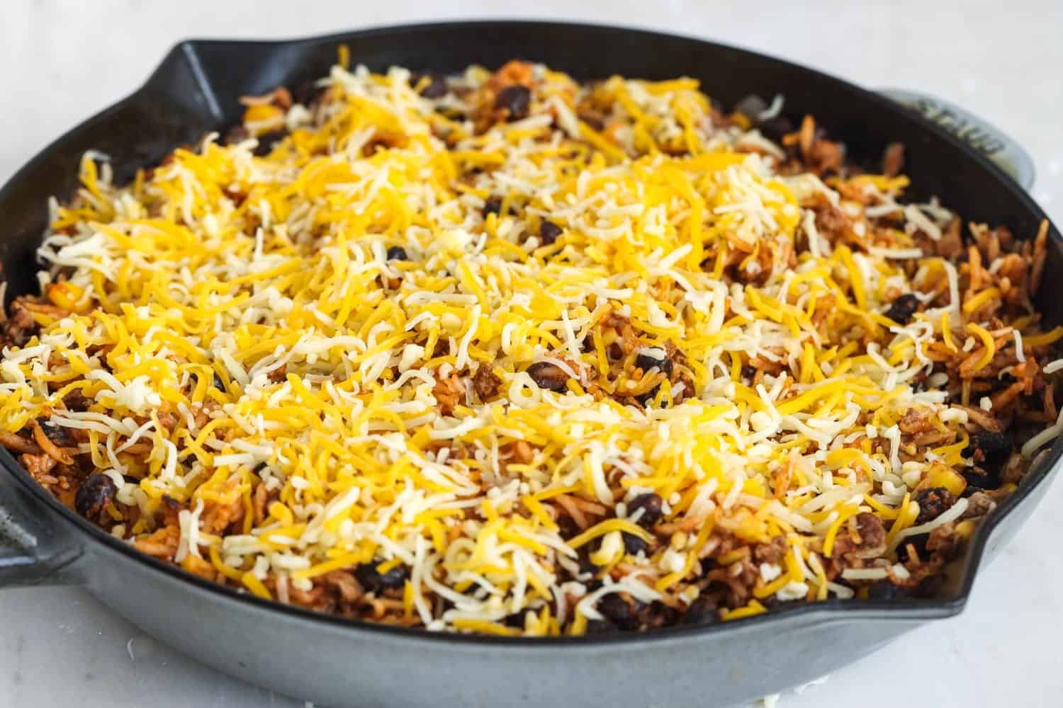 Shredded mexican blend cheese covering a skillet of cooked rice with beef and black beans.