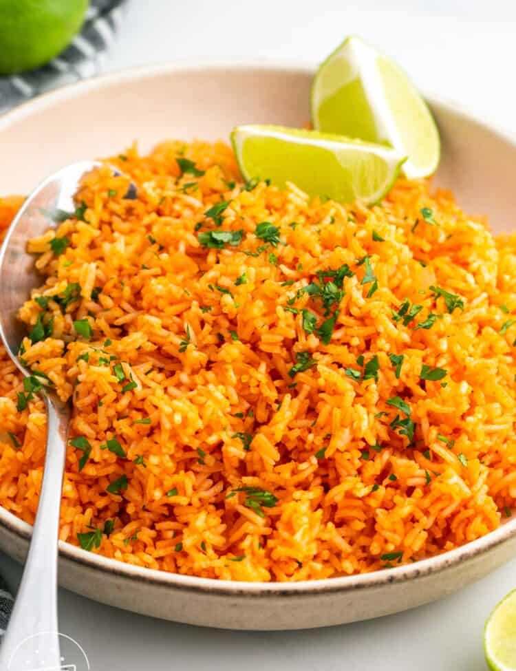 a white serving bowl of mexican seasoned rice. Lime wedges are on the side of the bowl, and a silver serving spoon is on the other side.