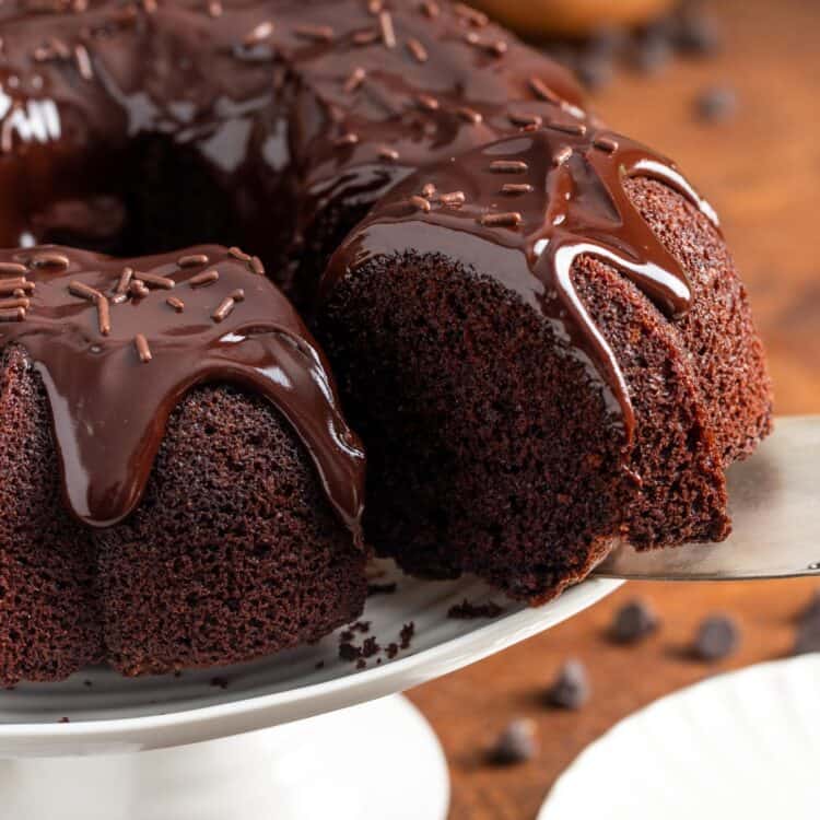a chocolate bundt cake with drippy chocolate ganache on a white cake pedestal. a spatula is removing a slice.