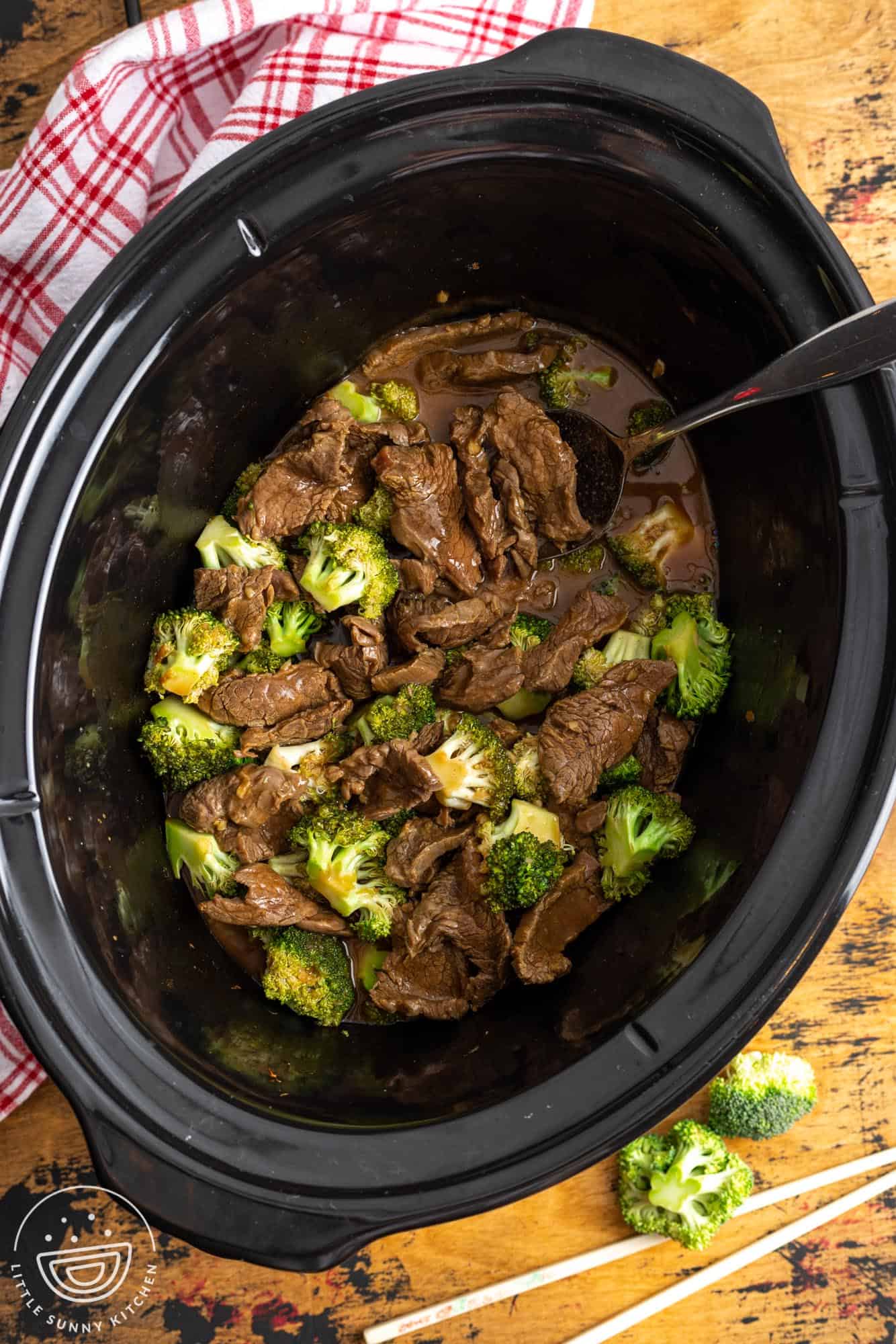 a black slow cooker on a wooden surface, filled with chinese beef and broccoli.