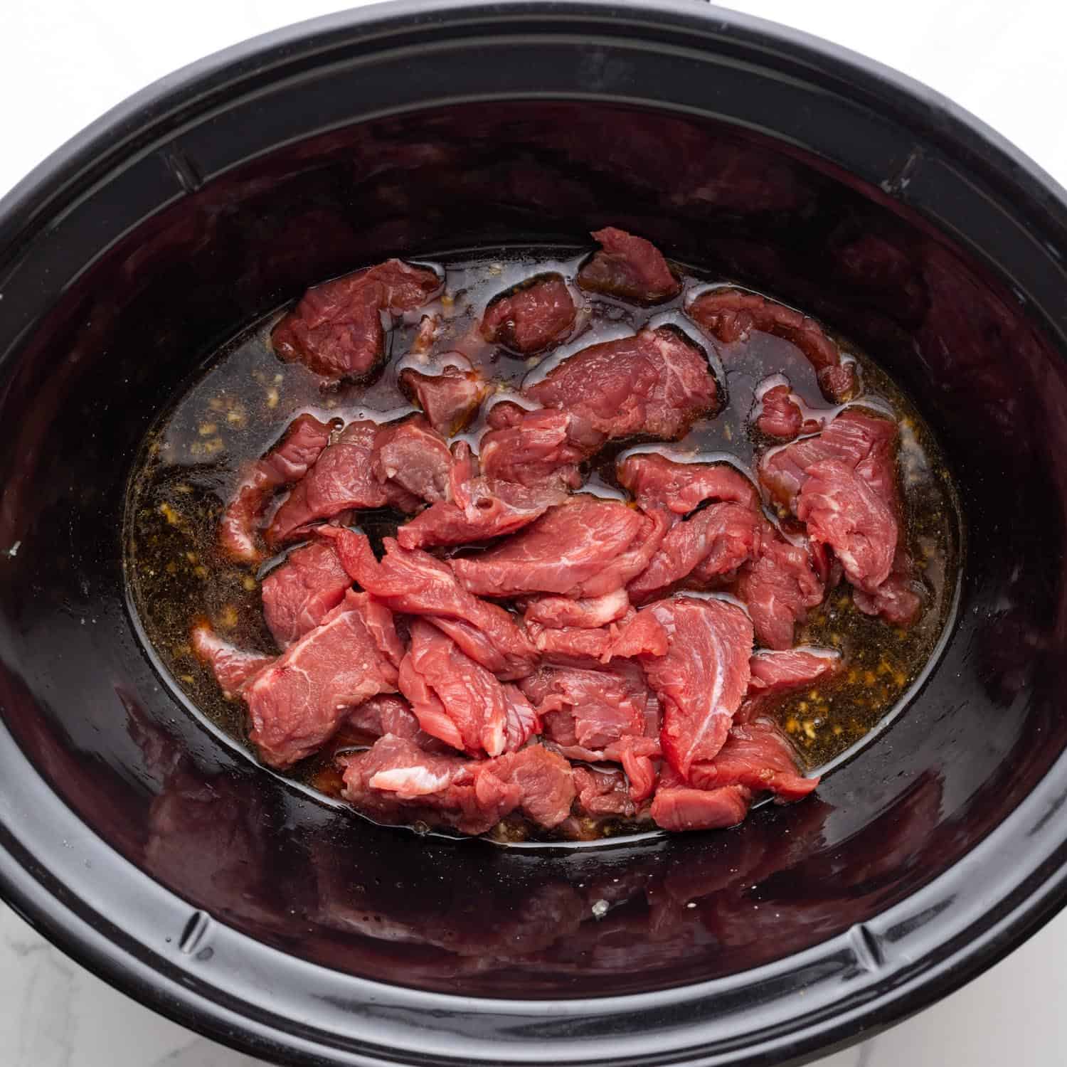 Sliced steak added to chinese inspired sauce in a black slow cooker pot.