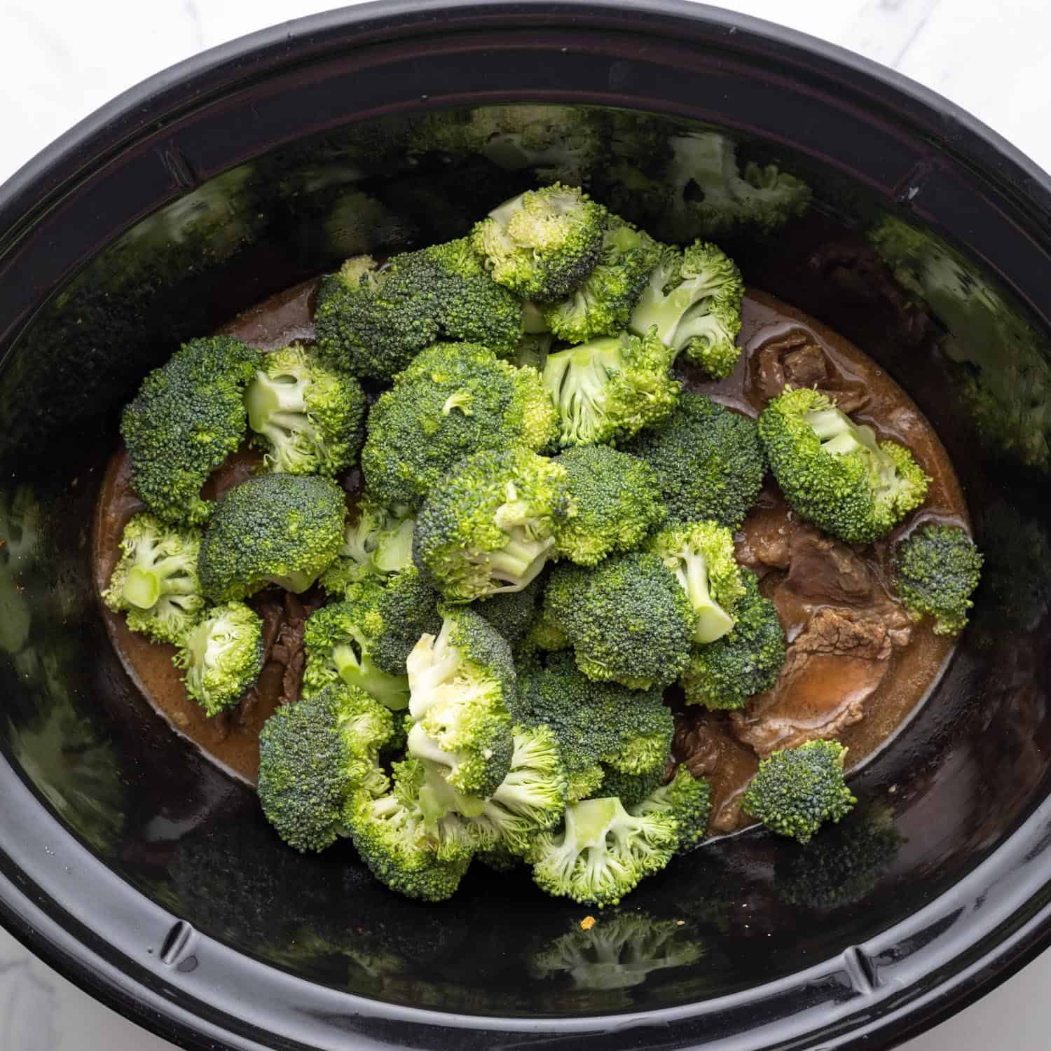 fresh broccoli added to a slow cooker filled with sliced beef and sauce.