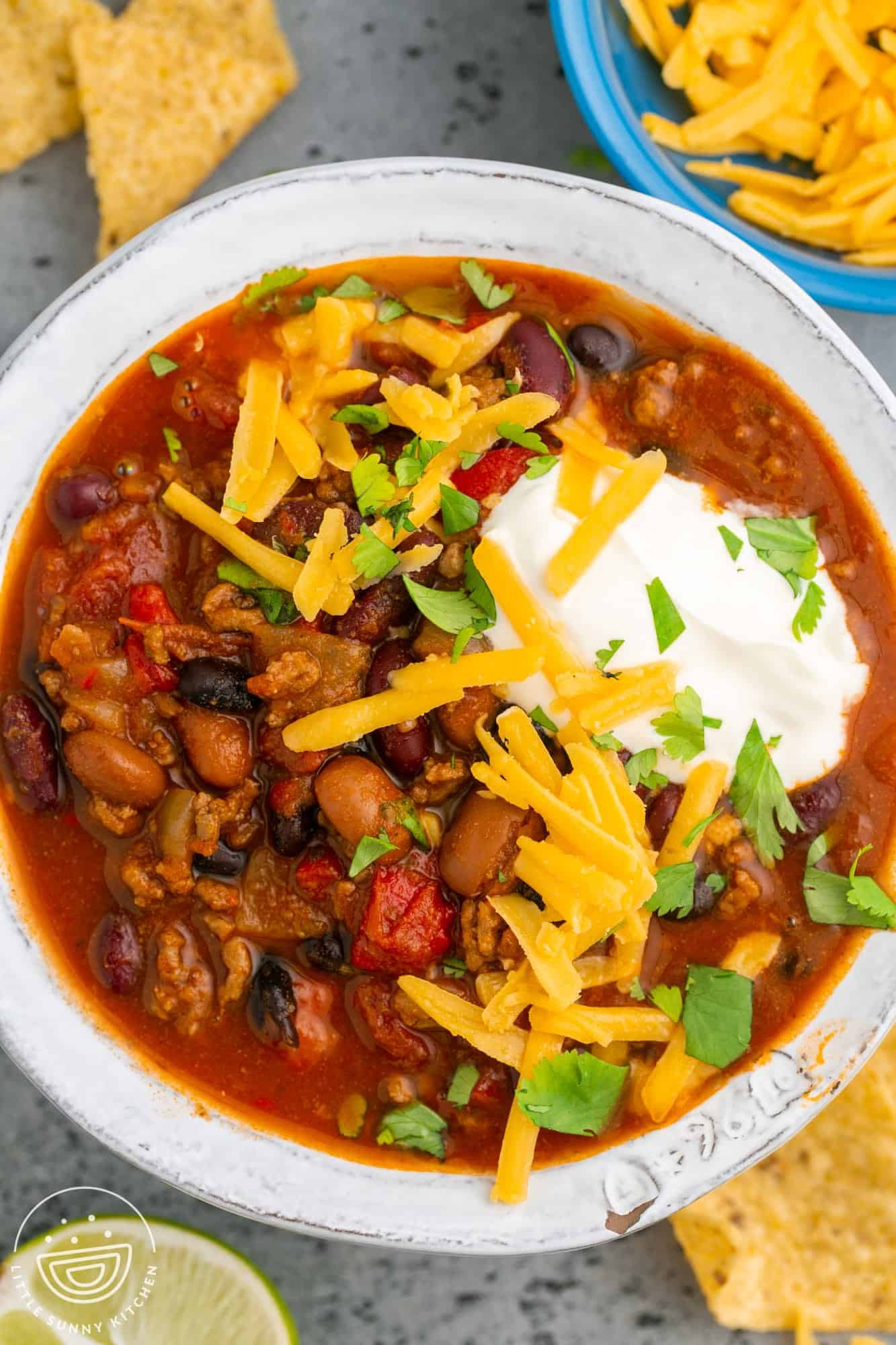 a white bowl filled with instant pot chili and topped with cheese and sour cream. Under the bowl are tortilla chips.
