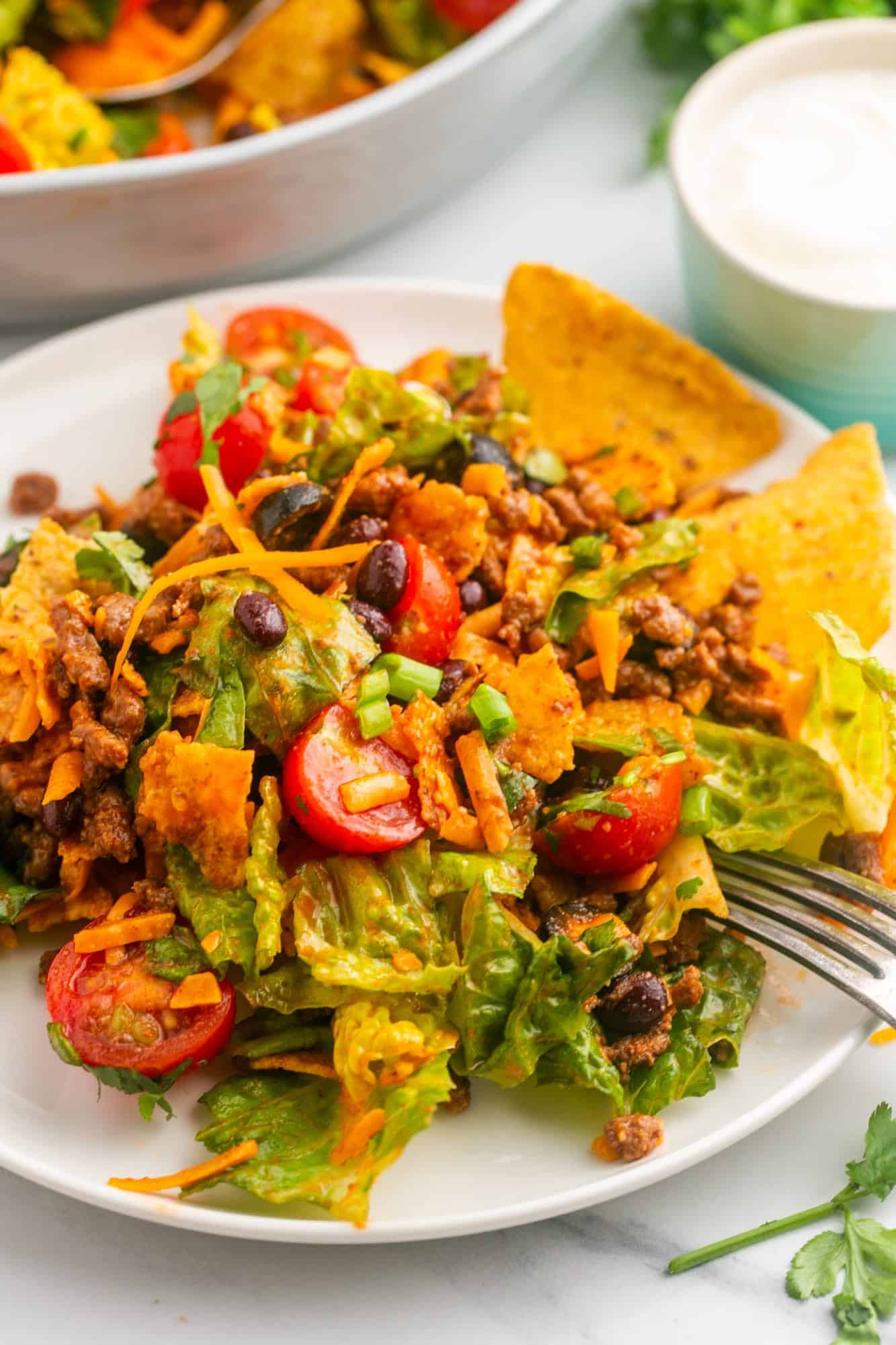 Dorito taco salad served on a white plate with a fork, and a small bowl of sour cream in the background
