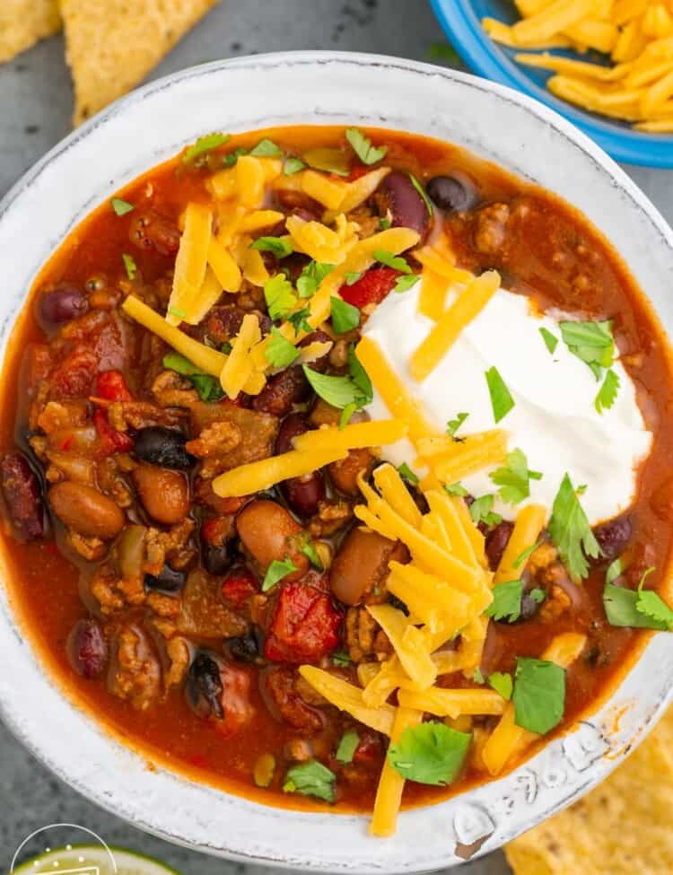 a white bowl filled with instant pot chili and topped with cheese and sour cream. Under the bowl are tortilla chips.