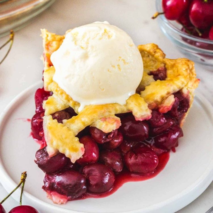 A slice of cherry pie served on a white dessert plate. Topped with vanilla ice cream.